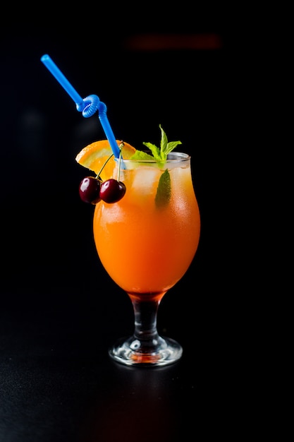 A glass of orange juice with cherries and mint in black background.