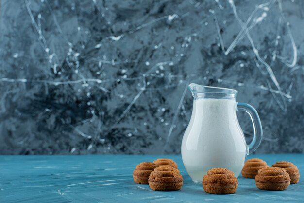 A glass pitcher of fresh milk with sweet round cookies on a blue background. 