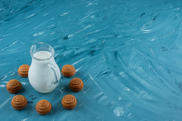 A glass pitcher of fresh milk with sweet round cookies on a blue background. 