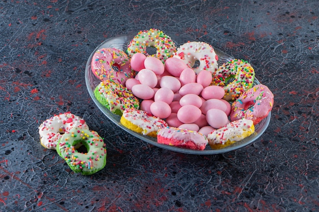 Стеклянная тарелка красочных вкусных пончиков и розовых конфет на темной поверхности.
