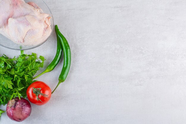 Glass plate of raw whole chicken with fresh vegetables on stone surface