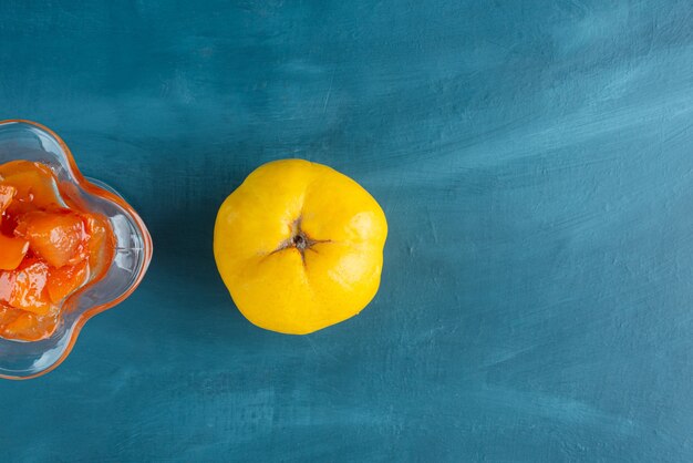 Glass of quince jam and quince fruit on blue surface.