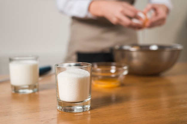 Glass of sugar at foreground with blur woman breaking egg in the bowl