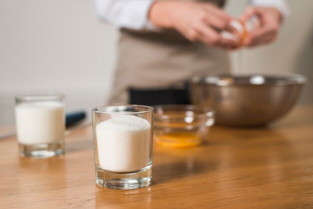 glass of sugar at foreground with blur woman breaking egg in the bowl