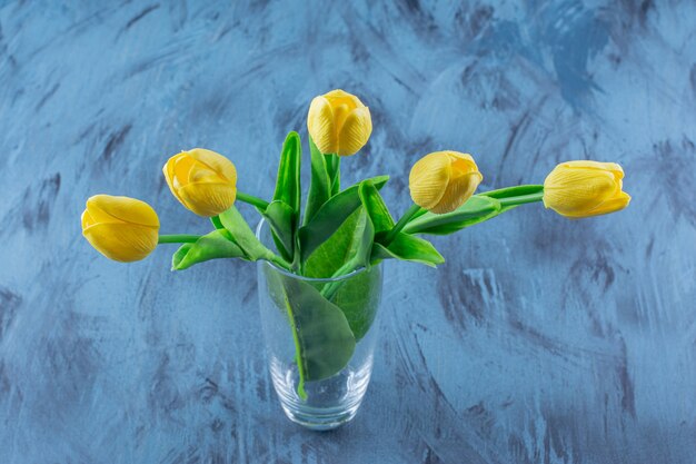 Glass vase of artificial yellow tulips on blue.