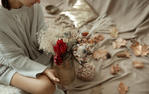Стеклянная ваза с осенними цветами в женских руках на размытом фоне с осенними листьями.