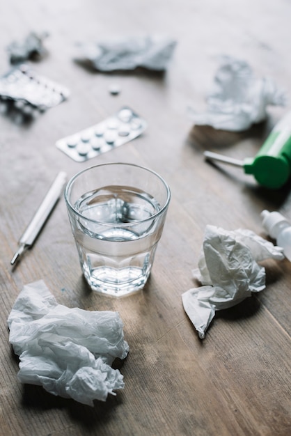 Glass of water; crumpled tissue paper; thermometer and medicines on wooden surface