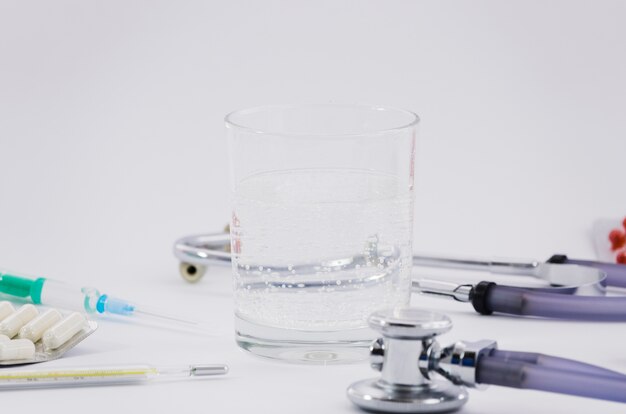 Glass of water; stethoscope; pills; syringe and thermometer on grey backdrop