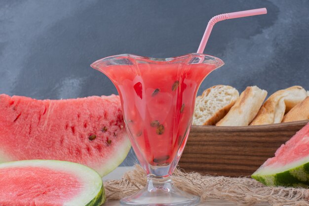 Glass of watermelon smoothie and basket of bread on white table.