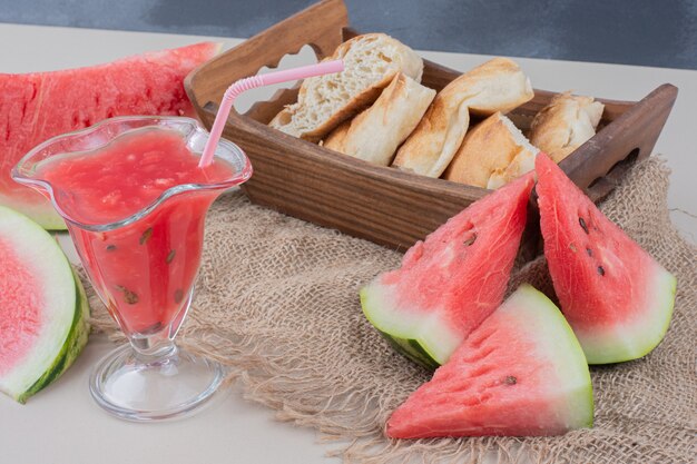 Glass of watermelon smoothie and basket of bread on white table.