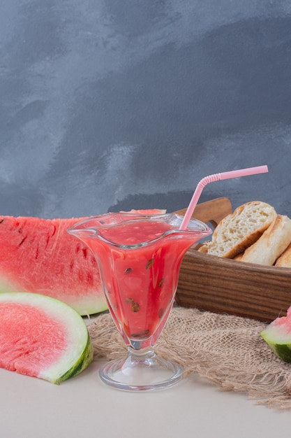 Glass of watermelon smoothie and basket of bread on white table.