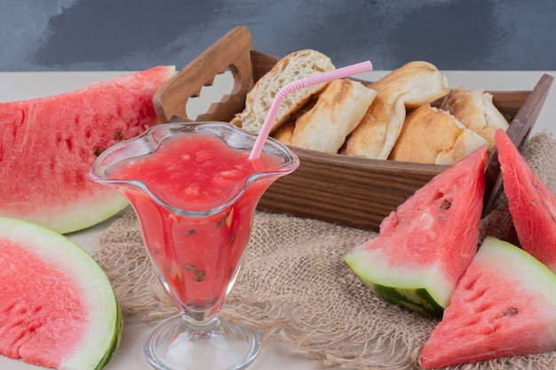 Glass of watermelon smoothie and basket of bread on white table.