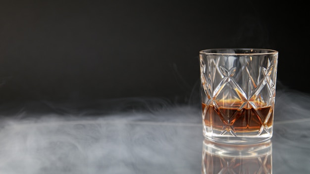 Glass of whiskey on a table surrounded by smoke against a black background