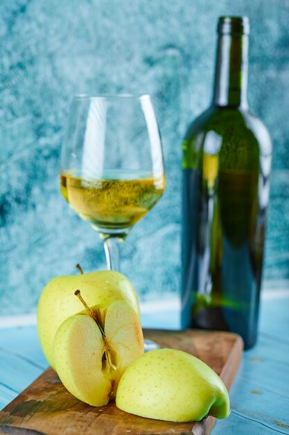 A glass of white wine and bottle with apple slices on blue wall.