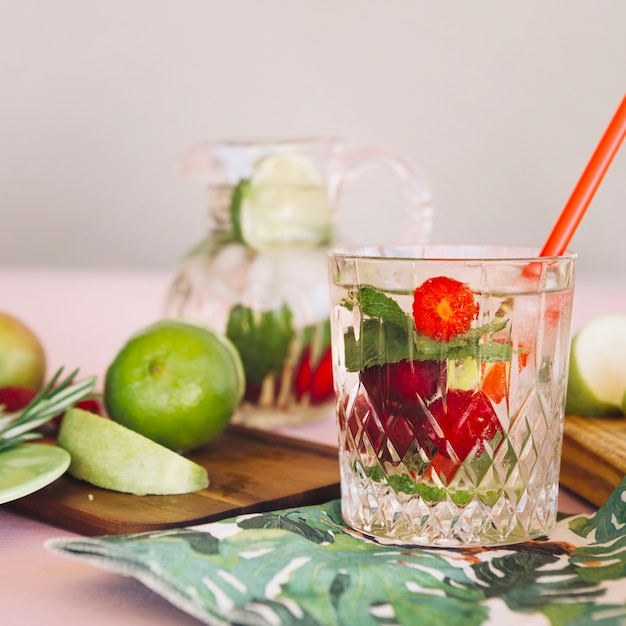 Glass with strawberries infused water near fruits on chopping board