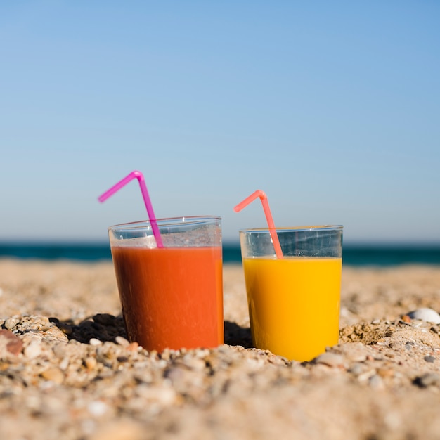 Glasses of orange and yellow juice with drinking straw on sand at beach against blue sky