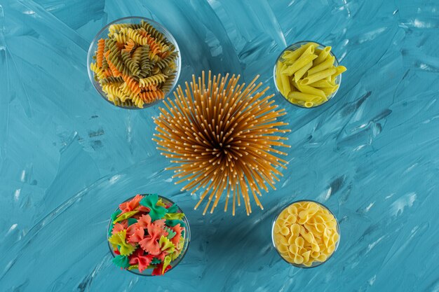 Glasses of various dry uncooked pasta on blue background. 