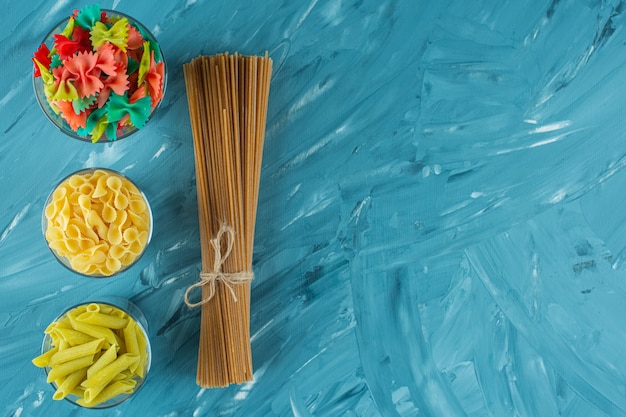 Glasses of various dry uncooked pasta on blue background. 