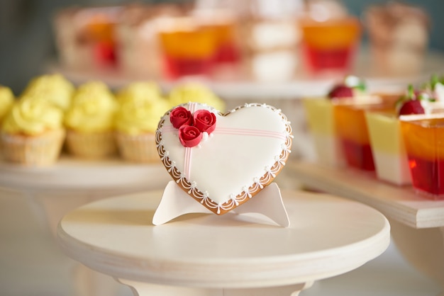 Glazed gingerbread heart stands on the wooden stand in the restaurant. It s decorated with sweet red roses and a little pattern. There is a colorful candy bar with cupcakes and jellies behind it.