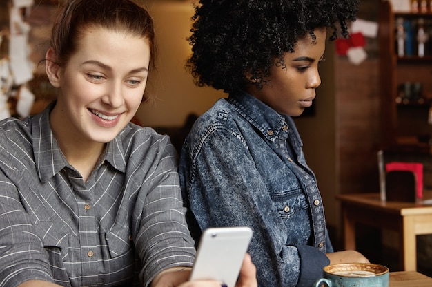 Gloomy African-American female having bored look, annoyed with her happy and cheerful Caucasian girlfriend