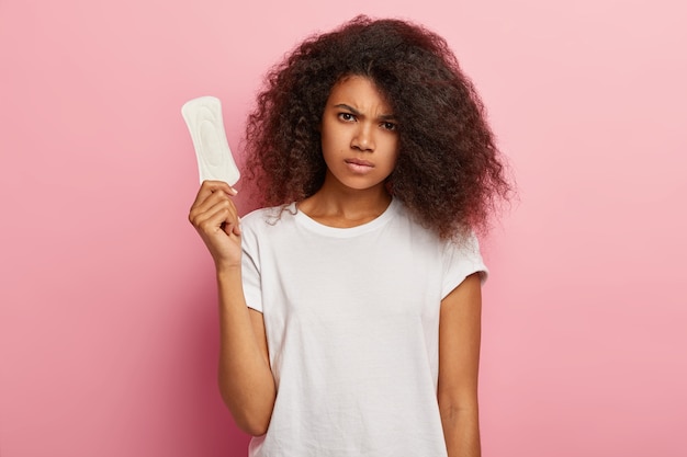 Gloomy African American woman cares about personal hygiene, holds sanitary pad