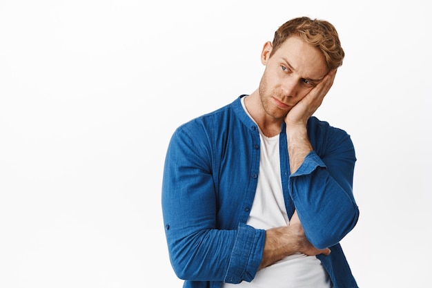 Gloomy and bored young redhead man, lean head on hand, looking aside with moody upset face, standing against white background