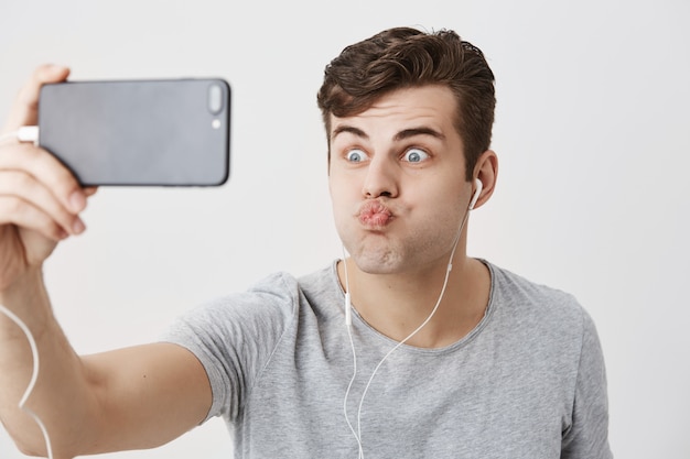 Gloomy caucasian male frowns face, holding smart phone in his hand, looking  with popped eyes, pouting lips, making faces, posing for selfie. Young good-looking guy having fun and mocking