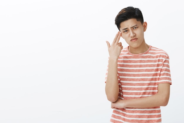 Gloomy cute asian guy suffering from boredom feeling pissed and irritated showing gun gesture near temple listeing eyebrow and grimacing from sorrow and boring feeling