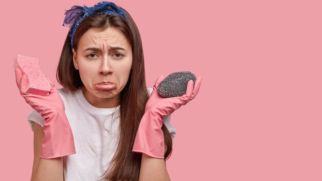 Gloomy displeased brunette woman purses lower lip, wears headband, feels upset as works long time on cleaning house