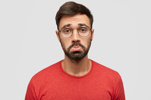 Free photo gloomy displeased sorrowful bearded young male purses lips in discontent, being offended by bad comments of followers, expresses negativity, wears red jacket, stands against white wall