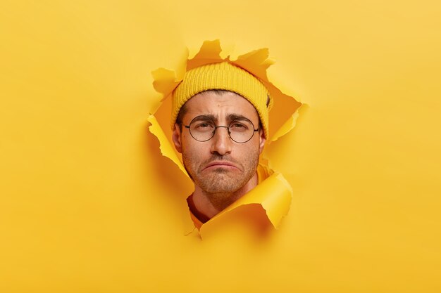 우울한 불만족 백인 남자는 부정적인 감정에서 얼굴을 능글 맞이하고 슬픈 표정을 지으며 노란 모자를 쓰고 있습니다.
