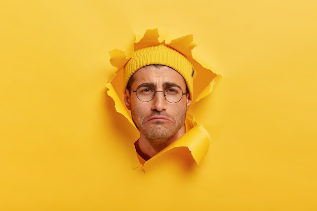 Gloomy dissatisfied Caucasian man smirks face from negative emotions, has sad look, wears yellow hat