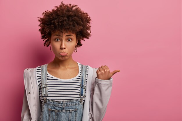 Gloomy insecure woman looks sadly and unhappily , points thumb aside, expresses regret as didnt get present for holiday, shows direction right, dressed in casual wear, isolated on pink wall