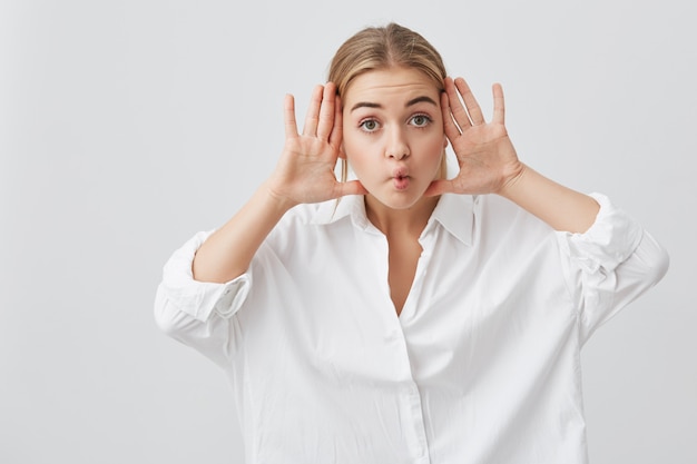 Gloomy, playful, having fun on camera beautiful caucasian female dressedin white shirt mocking at cemera. Pretty girl popping her eyes, holding palms open behind her ears, posing indoors.