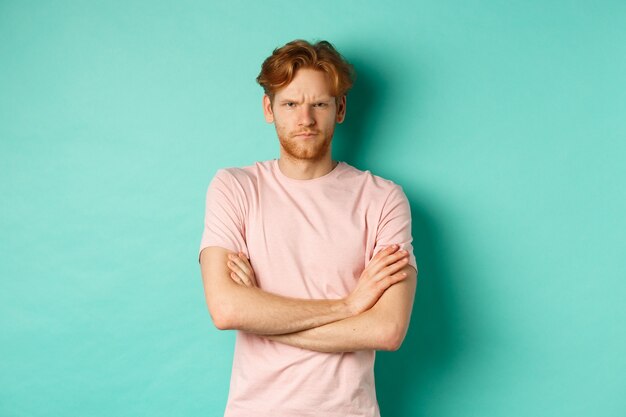 Gloomy redhead guy feeling offended, frowning upser with arms crossed on chest, looking insulted and sulking, standing over mint background