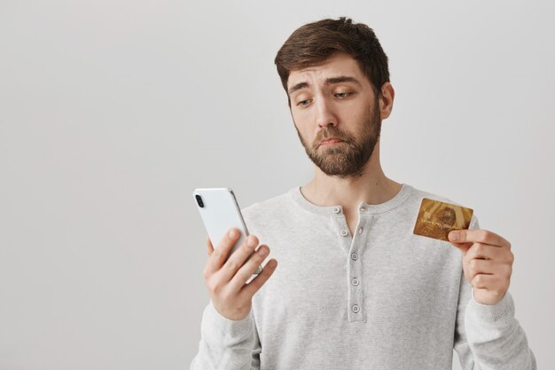 Мрачный и грустный парень смотрит на смартфон, держа кредитную карту