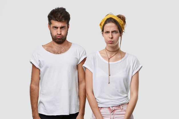 Бесплатное фото Мрачные угрюмые женщина и мужчина сосут щеку, не разговаривают друг с другом после ссоры, имеют разногласия, оскорблены плохими словами, стоят плечом к плечу в помещении, носят белую повседневную футболку-макет