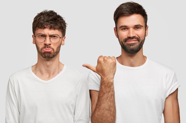 Free photo gloomy unshaven young man model with grumpy expression, being in low spirit, stands near best man companion, wear casual white t shirts, express different emotions