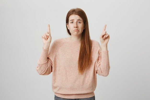 Gloomy upset redhead girl pouting indecisive, pointing fingers up