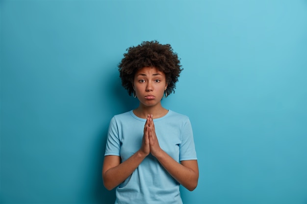 Gloomy upset woman with Afro hair keeps palms pressed together in pray, supplicates over blue wall, needs your help, begs for favor, wears casual t shirt, makes innocent face expression