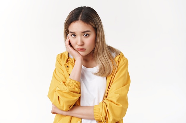 Free photo gloomy young sad asian blond girl sighing upset stoop look from under forehead lonely unhappy lean palm bored feel regret sadness moody expression standing white wall pessimistic