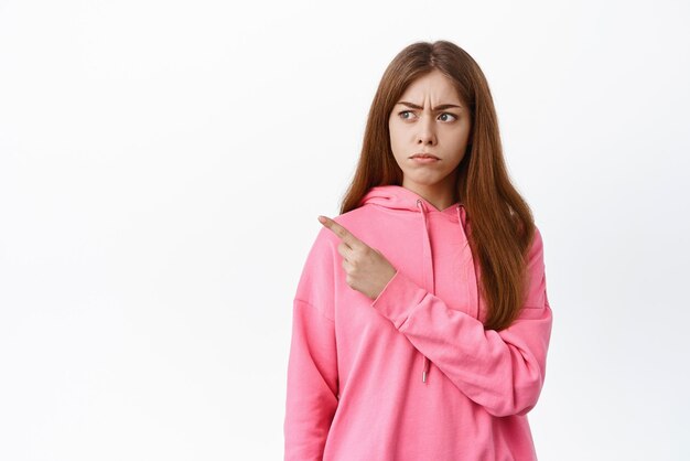 Gloomy young woman frowning and looking displeased aside points left with disapproval dislike something bad standing over white background