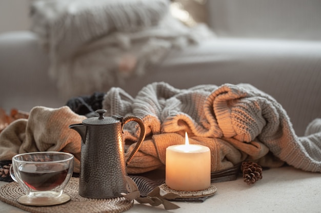 A glowing candle, a cup of tea and a teapot against the space of warm sweaters in the room.