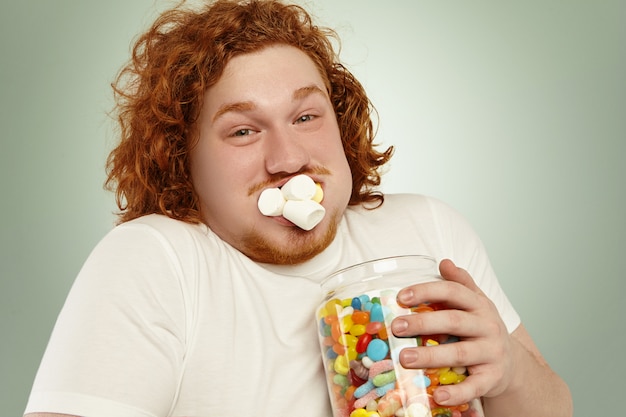 Gluttony, obesity, junk food and unhealthy lifestyle. Funny redhead obese young man holding big jar of sweets and marmalades, having happy and joyful expression, his mouth full of marshmallow