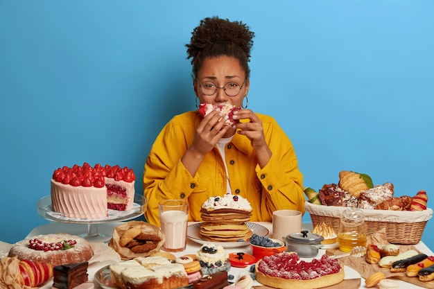 Free photo gluttony and overeating concept. upset crying ethnic woman eats piece of cake reluctantly, sits at table with many desserts, isolated over blue wall