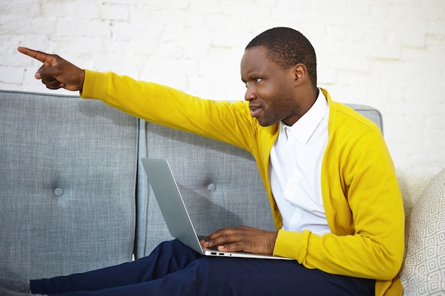 Go away. Sideways shot of attractive young dark skinned man having angry look, being disturbed by someone while watching movie online on laptop, sitting on couch and pointing finger at door