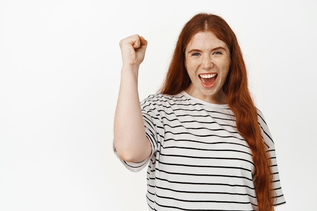 Go team. Cheerful young redhead girl, female activist clench fist and pump arm up to chant, rooting for someone, marching for human rights and unity, standing against white background.