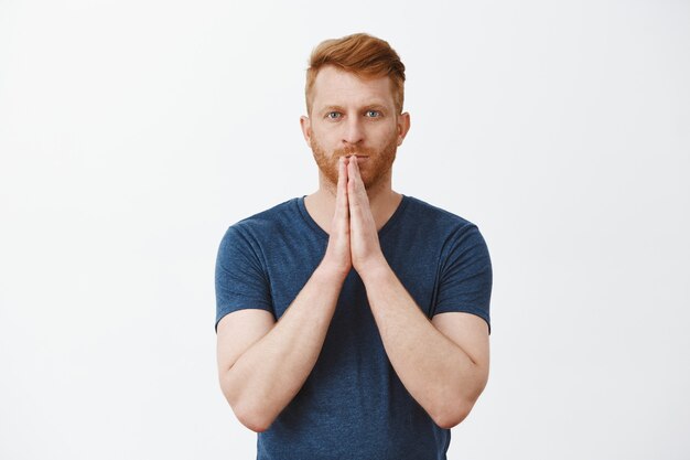 God please hear my prayers. Determined and focused attractive male redhead entrepreneur being worried for business, holding hands in pray over lips and staring patiently, waiting for miracle