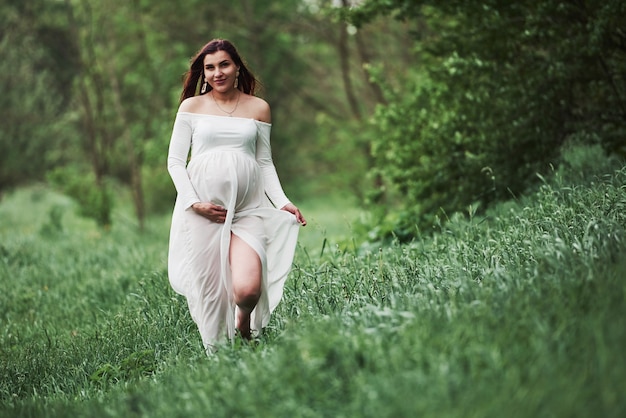 Going forward and smiling. Beautiful pregnant woman in dress have a walk outdoors. Positive brunette