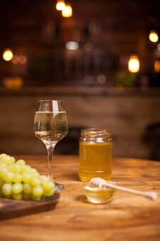 Golden honey jar next to a refined glass of white wine over a rustic table in a vintage pub. wine degustation. delicious grapes.
